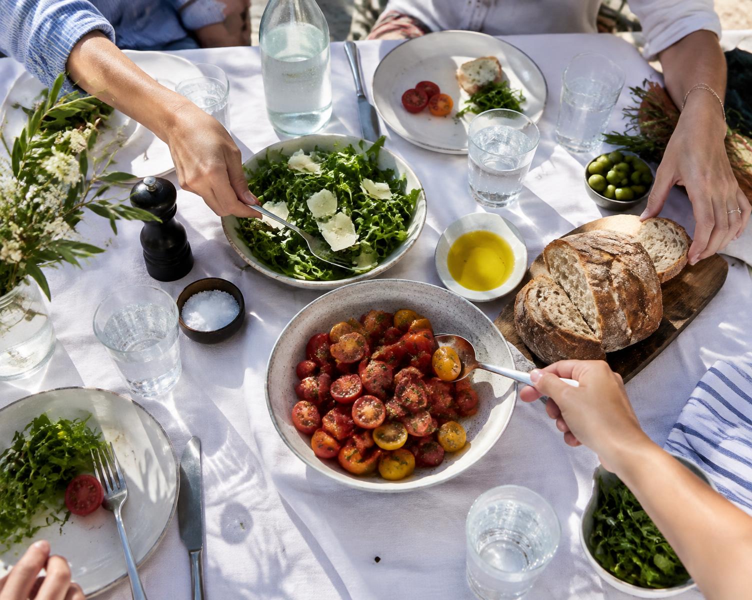 Hands reaching across an outdoor table of tomatoes, greens and bread
